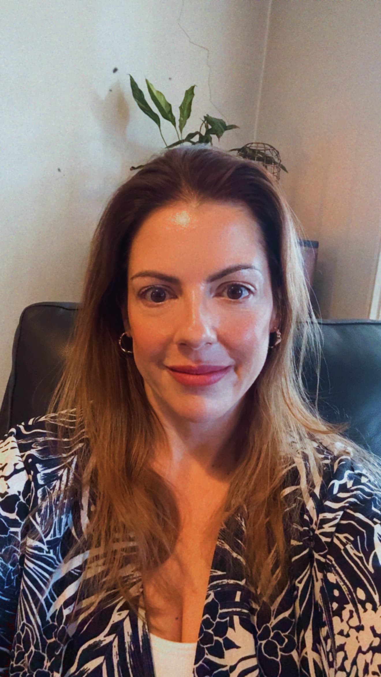 Smiling woman with long brown hair wearing a black and white patterned top indoors.