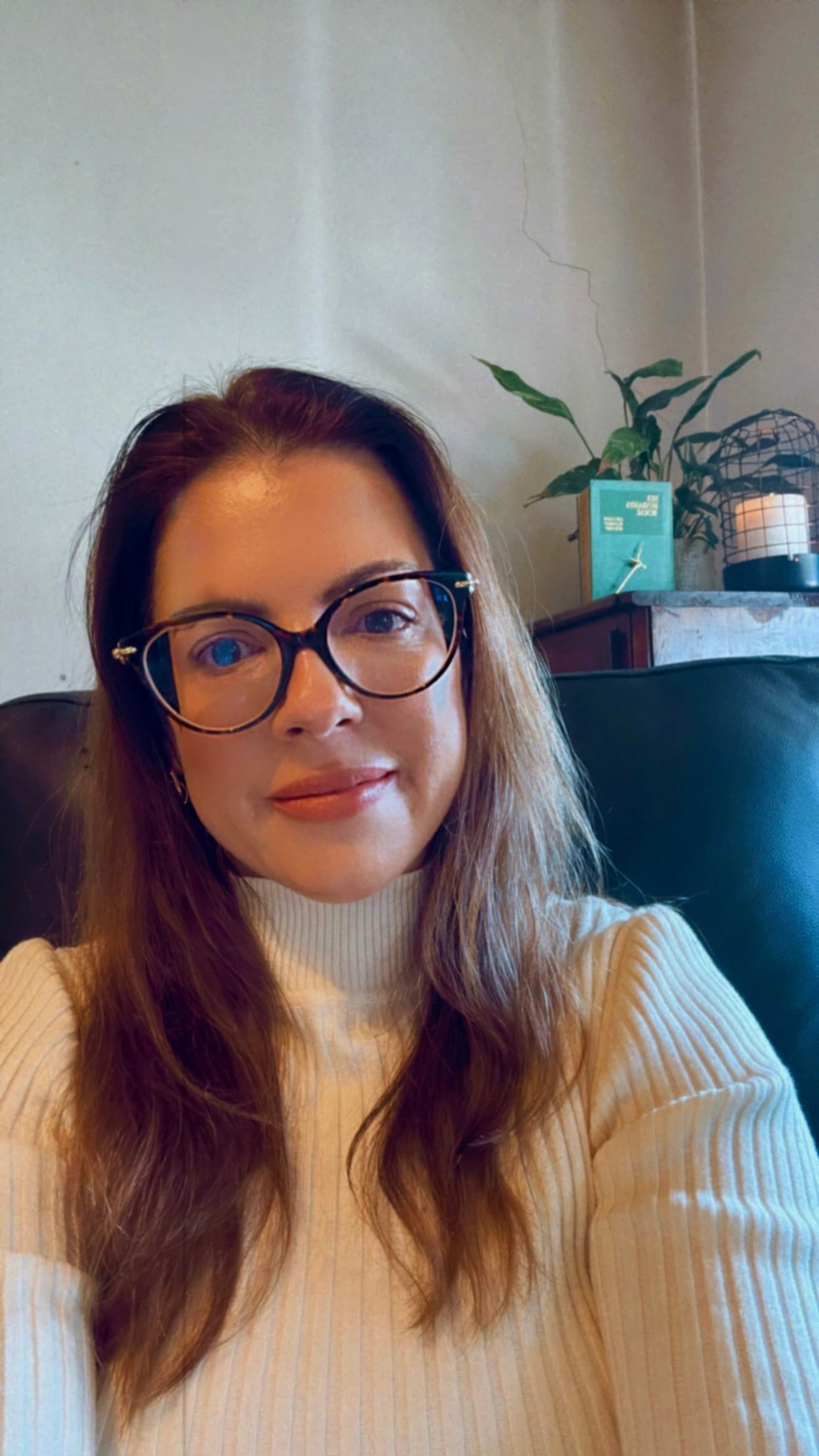 Woman with glasses and long brown hair wearing a white ribbed turtleneck sweater indoors.